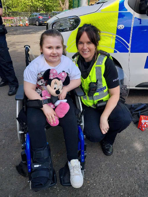 The image shows a young girl who is sitting in a wheelchair next to a female police officer who has full uniform. The Police Officer is crouched down beside the young girl and both are smiling. 