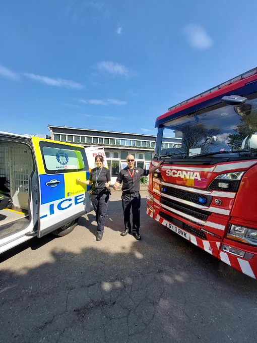 The image shows a Police vehicle and a Fire engine parked together. In between the vehicles is a female police officer and a male fire fighter who have handcuffed them selves to each other attempting to show "joint up" working. 
