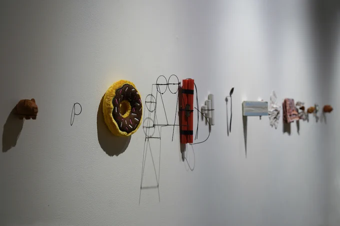 A photograph of a series of objects hanging on a white gallery wall. Objects include a stuffed doughnut, a silver spoon, dynamite, wire sculptures, and more. 