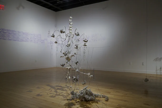 A photograph of a gallery installation. Objects hanging from the ceiling fill the space. The objects are in shades of blue, white, and silver and hang in rows on thin wires. A chandelier caught in objects hangs close to the ground in the foreground of the image. 
