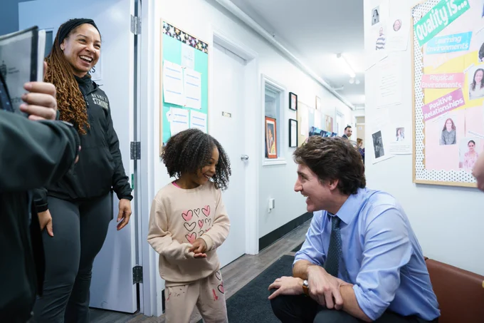 Prime Minister Justin Trudeau crouches down to smile at a young girl at a child care facility, who is smiling at him. Her mother watches on.