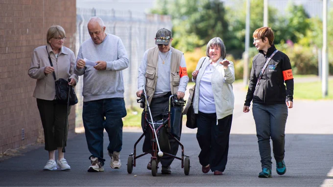 A small group of people, including one using walking aids, talk and smile as they enjoy a guided Health Walk along a path.