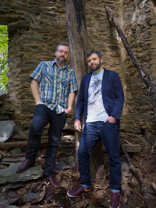 Night Vale creators Jeffrey Cranor and Joseph Fink standing outside, near ruins. Jeffrey is wearing red glasses, a light green and blue plaid shirt and dark jeans. Joseph is wearing a navy blue suit jacket over a t-shirt and dark jeans. A stone wall rises behind them.
