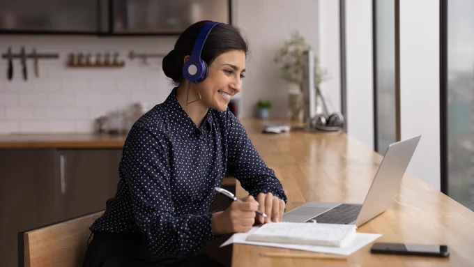 A woman is sitting at a table, wearing a headset and looking at a table while holding a pen. A kitchen is visible in the background.