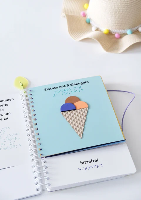 Aufgeschlagenes Ringbuch mit einer tastbaren Illustration (Eistüte mit drei Eiskugeln), darunter das Wort "hitzefrei" in Braille und Großdruck, oben rechts über dem Buch ein Teil eines Sonnenhutes
