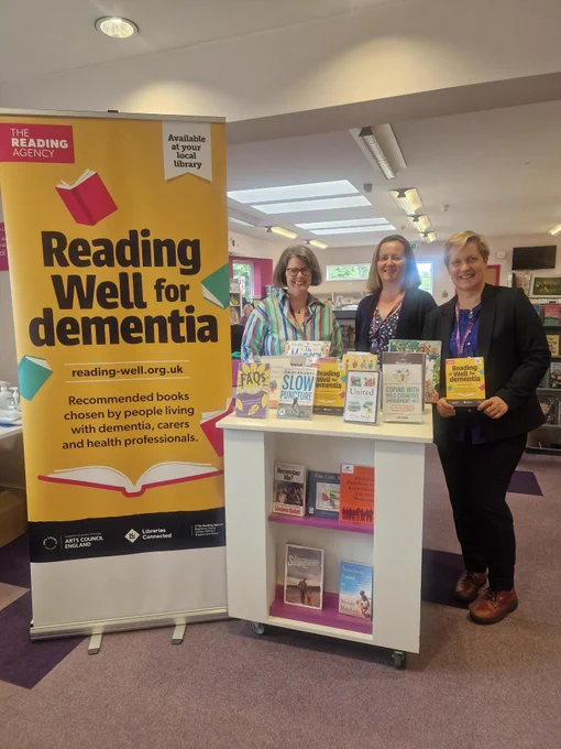 Banner reading well for dementia. Display cabinet of books . three ladies standing behind the cabinet of books.