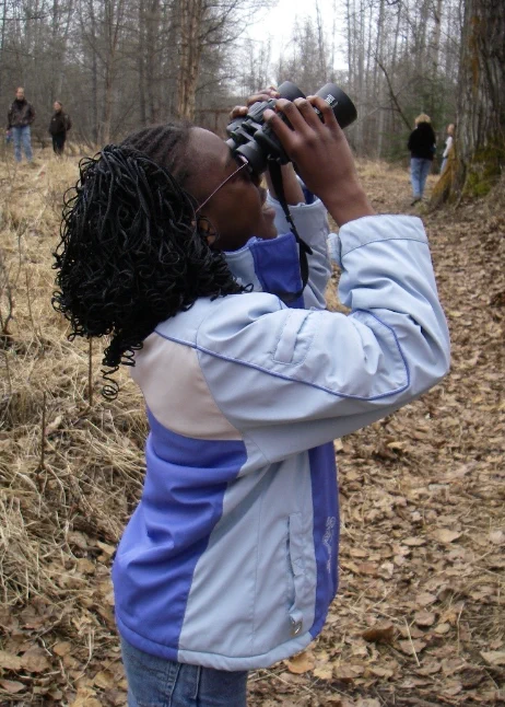 Photo caption: Spring is a great time to view and learn about birds! Billions of birds return to Alaska for the summer. 
 Photo by BLM.  
 A student using a pair of binoculars, looking up for birds. 