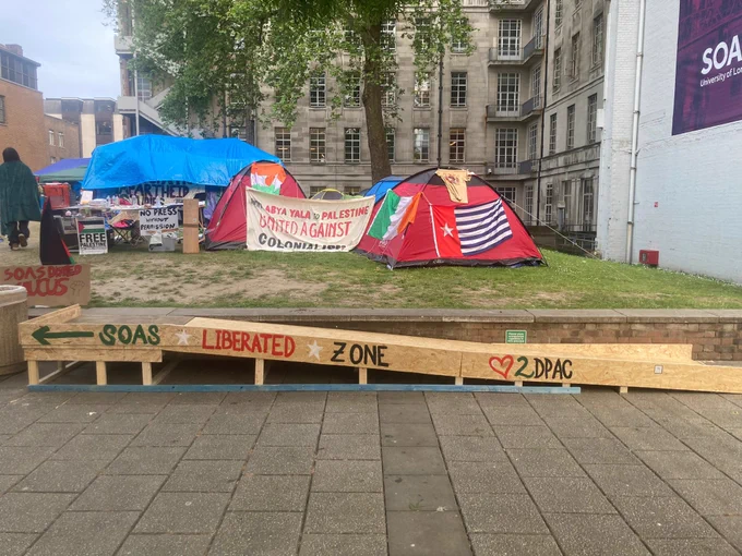 Wooden wheelchair ramp adjacent to a wall supporting a raised green containing a protest camp. Hand painted along the edge of the ramp, in Palestinian flag colours are the words “SOAS Liberated Zone”. Further down the edge of the ramp is a hand painted image of a love heart follow by a 2, then DPAC. In the background on the green are various tents and banners. In the foreground is a section of pavement.