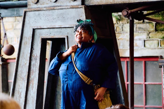 Cherie as Miriam in a blue boiler suit, satchel and headscarf.