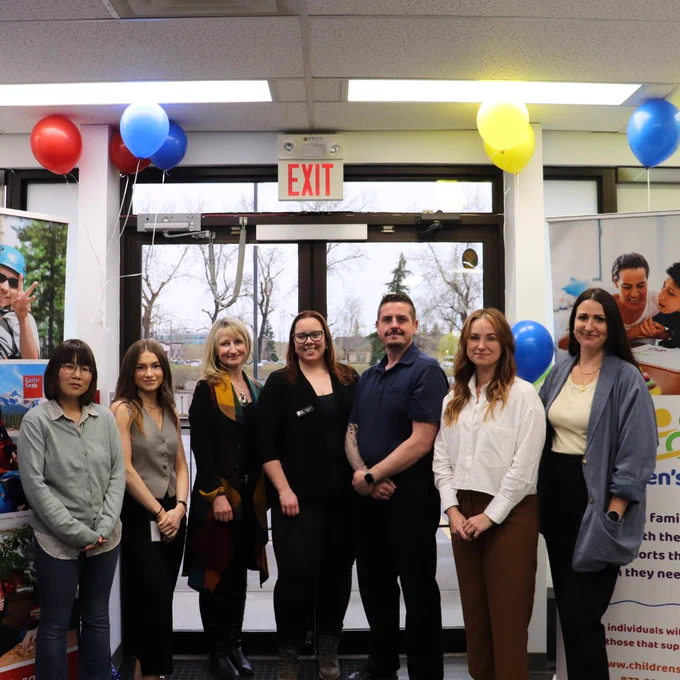 Seven members of the Easter Seals Alberta leadership stand together and are smiling at the camera. They are standing in the Easter Seals Alberta office at an open house. The Easter Seals Alberta banner is behind everyone on the left side of the image and the Children's Link banner is behind everyone on the right side of the image. There are seven red, yellow, and blue balloons behind them.