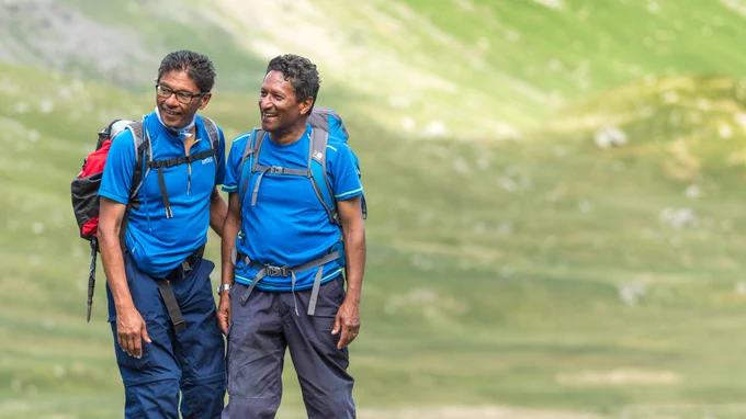 Two people smiling and looking to the left of the image. They are wearing walking clothes and rucksacks, the background is a green, rolling hillside.
