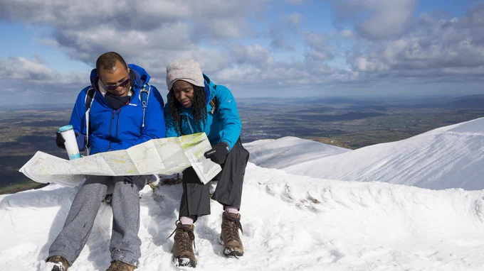 Two people are sat on a snowy hill top, inspecting a map.