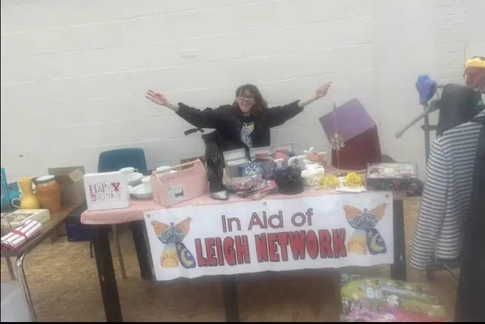 Image: Faye wearing a Leigh network T-shirt with arms spread wide showing the stall with lots of different items on sale.