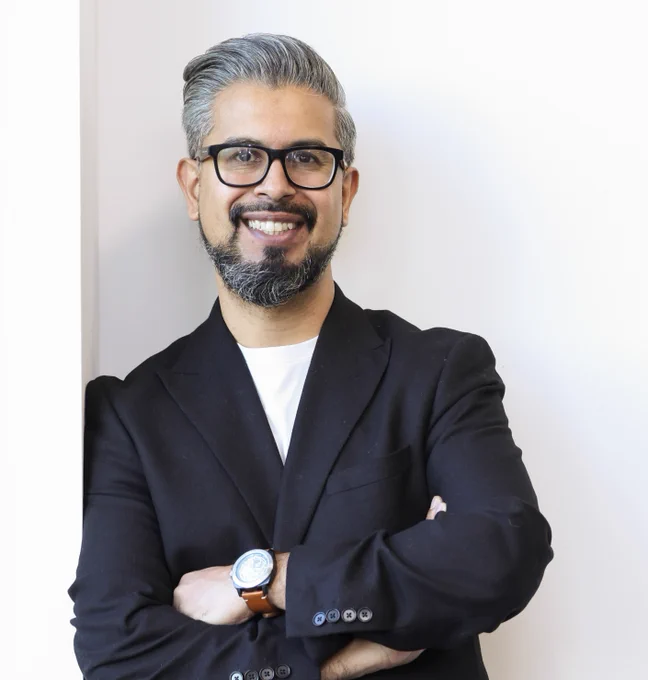 A man is leaning against a wall. He is wearing glasses and a navy blazer. He is smiling at the camera.