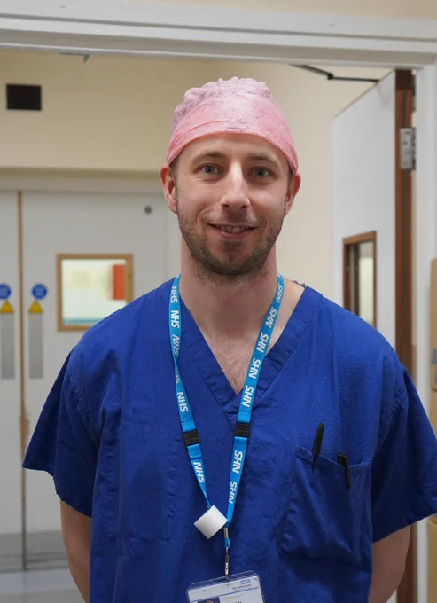 Sean smiles into the camera, he's wearing blue surgical scrubs, an NHS lanyard, and a hair cap. 