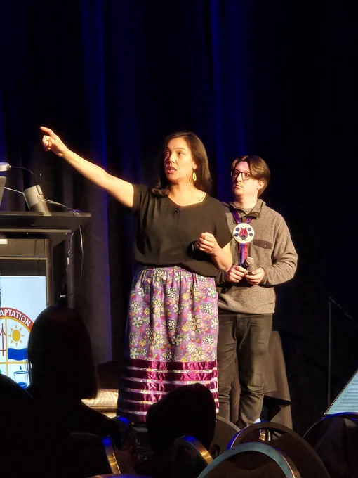 Photo of Bazile Panek & Ziigwanikwe Katy Bresette on stage next to a podium. Katy is pointing to the side while speaking. 