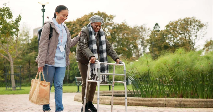 a person holding a tote bag assists another person with a walker walk through a park. 