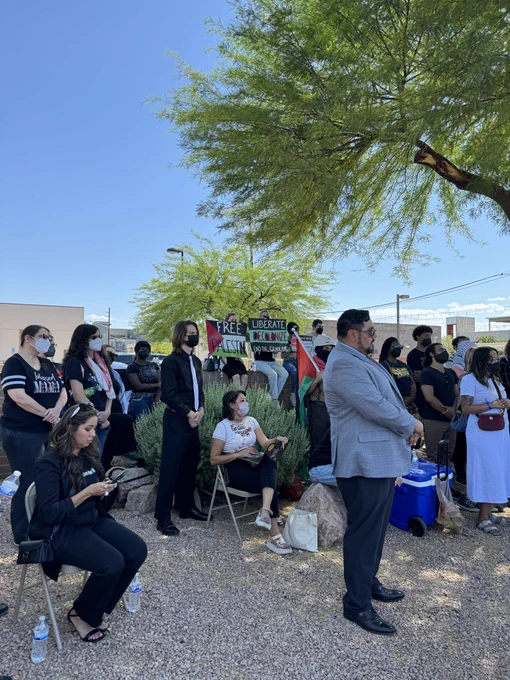 Community members, incl. students, an attorney & the family of folks arrested, gather around a press conference hosted after an arraignment hearing on May 14, 2024. People seated on a brick wall in the back hold protest signs.