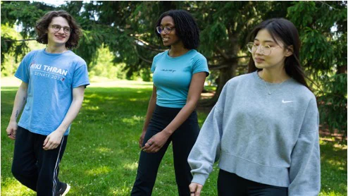 Three people walk together outside in a grassy area with trees in the background.