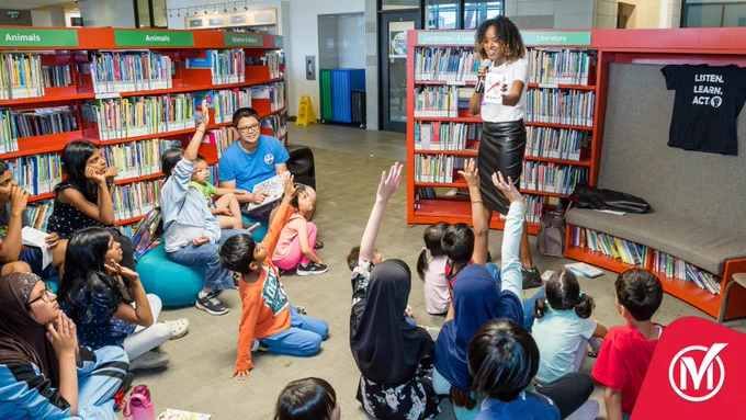 Group of children at the library, surrounded by books, while listening to a speaker reading a book. 