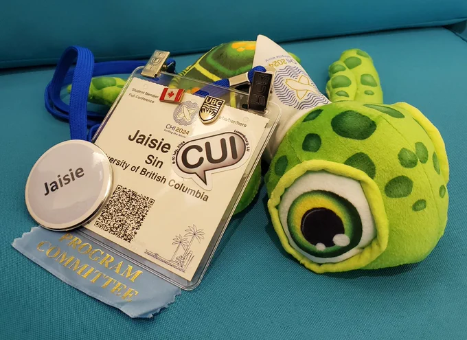 Jaisie's CHI 2024 name tag leaning on a CHI turtle plushie. Attached to the name tag are a "Program Committee" ribbon, a "CUI" conference sticker, a Canada flag pin, and a UBC pin. The turtle plushie is wearing a scarf labelled "CHI 2024" and has the conference logo.