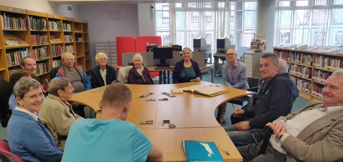 a large table with 13 of our service users and volunteers all smiling. Craig who works for the library is sitting with a large archive record book in front of him 
