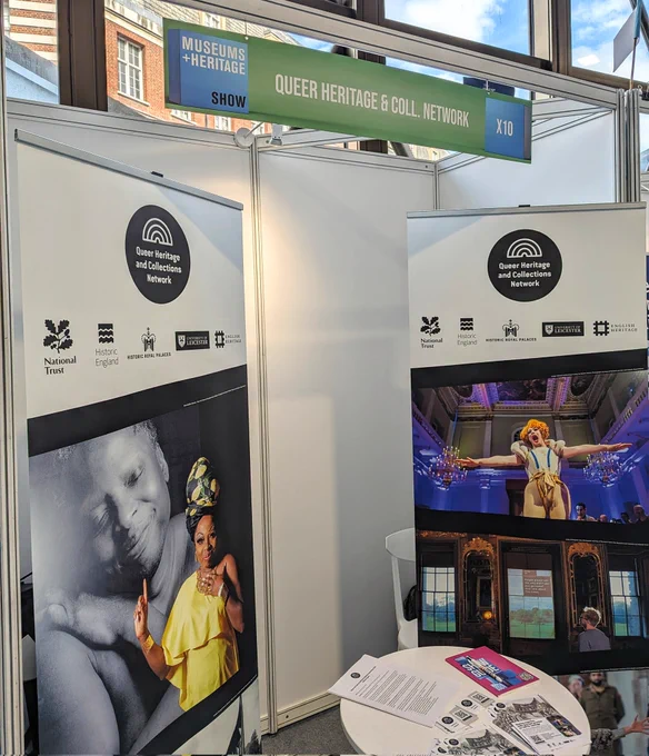 Convention stand with two promotional banners with the Queer Heritage and Collections Network logo and photos of performers in heritage places.