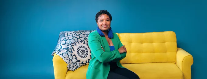 Terresa Moses wearing bright blue earrings and a vibrant green cardigan sitting on a sunny yellow couch