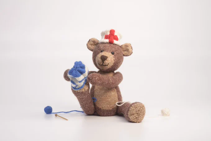 A knitted toy bear wearing a nurse's hat putting on a half-finished woolen sock on their foot. 