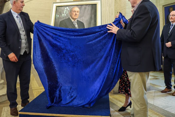 The picture shows the current Assembly Speaker Edwin Poots MLA (left) and the former Assembly Speaker Mitchel McLaughlin (right) remove a blue velvet cover to reveal the portrait of the former Speaker. The portrait sits on a easel upon a blue riser. In the background is the marble Great Hall staircase with its intricate bronze balustrade and a lifesize sculture of Lord Craigavon sits at the top of the staircase.   