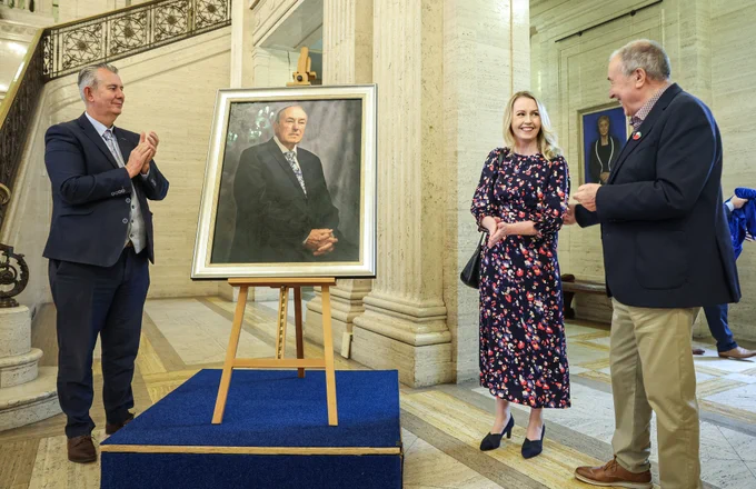 The picture shows the current Assembly Speaker Edwin Poots MLA (left) clapping and looking at the portrait. The Artist Catherine Creaney and former Assembly Speaker Mitchel McLaughlin  are both stood on the right of the portrait, they are looking at each other and smiling.) The portrait sits on a easel upon a blue riser. In the background is the marble Great Hall staircase with its intricate bronze balustrade and a lifesize sculture of Lord Craigavon sits at the top of the staircase.   
