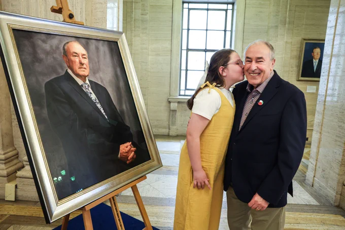 The picture shows the former Assembly Speaker Mitchel McLaughlin receiving a peck on the cheek from his  grandaughter. He is looking towards the camera and smiling. The portrait of the former Speaker sits on a easel upon a blue riser to the left of the image. The portrait of the Former First Minister David Trimble can be glimpsed on a marble wall panel in the background.  