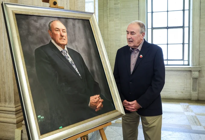 The picture shows the former Assembly Speaker Mitchel McLaughlin looking at his portrait, his hands are clasped reflecting how his hands are clasped in the portrait. The portrait of the former Speaker sits on a easel upon a blue riser to the left of the image. 