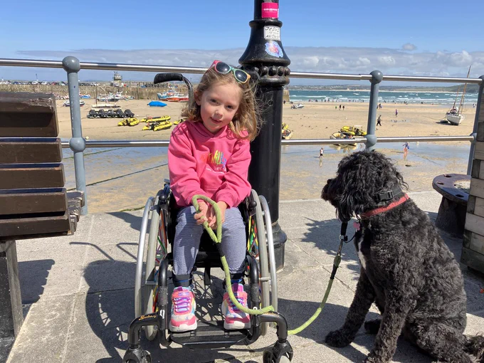 A photo of a young girl in a pink jumper in a wheelchair. She is holding the lead of her black dog and is at the seaside.