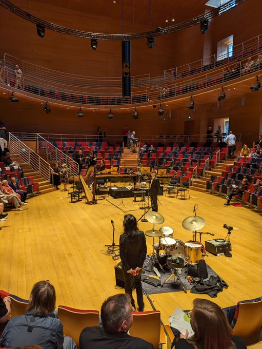 This is a photo of the Pierre Boulez Saal, a concert hall in Berlin. It is a large round room with a stage that is lower than the seating. The seating is raked, and surrounds the stage on all sides. The flooring is a light polished wood, and there is a drum kit and harp visible, among other instruments.