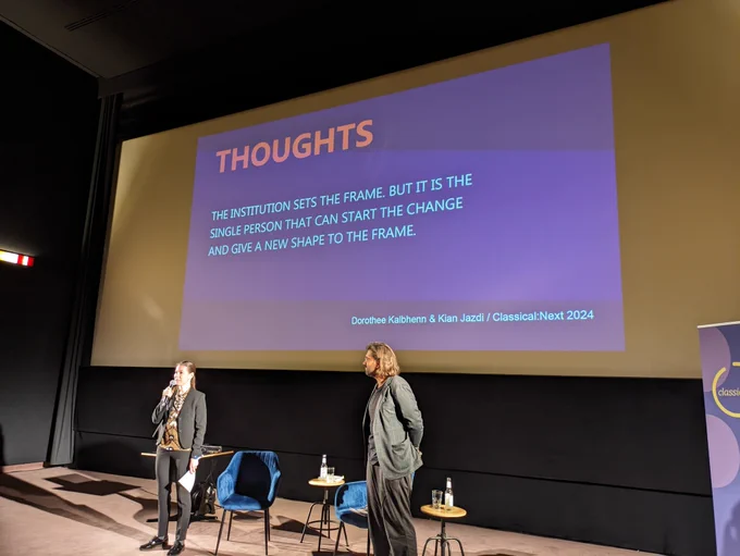 This is a photo of Dorothee Kalbhenn and Kian Jadzi delivering a talk in front of a projection screen. The screen reads "Thoughts: The institution sets the frame. But it is the single person that can start the change and give a new shape to the frame"