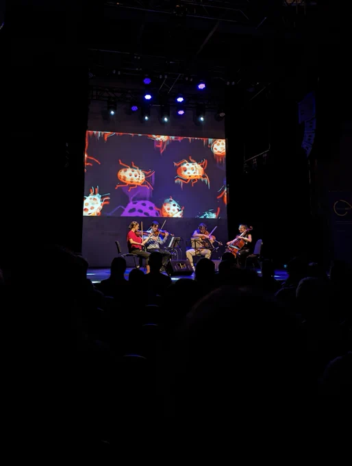 This is a photo of the Ligeti Quartet performing on stage, taken from the audience perspective. The stage is lit, and behind and above the performers there is a projection screen. The projection screen has round shaped orange cartoon dotted insects on it. 