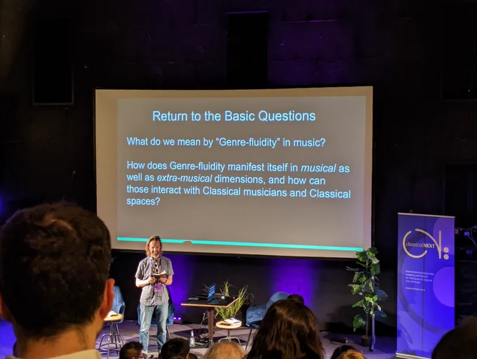 This is a photo of Gabriel Prokofiev in front of a projection screen, which reads: "Return to the Basic Questions: What do we mean by Genre-fluidity in music? How does Genre-fluidity manifest itself in musical as well as extra-musical dimensions, and how can those interact with Classical musicians and Classical spaces?