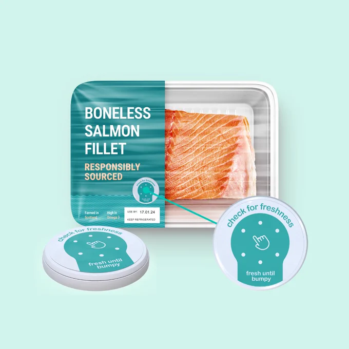Against a soft green backdrop, the new circular-shaped Bump Tag is attached on a pack of salmon fillet beside its expiry date. Below, the Bump Tag is enlarged in two perspectives—a side view and a top view—with curved text on the tag urging: “Check for Freshness” and “Fresh Until Bumpy.”