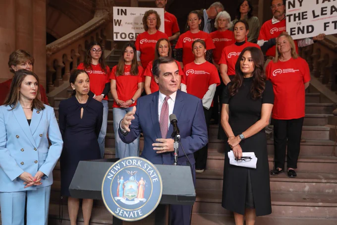 Senator Gianaris wears a navy suit and light red tie as he speaks at the podium. Behind him, advocates wearing red t shirts are standing on the steps of the Million Dollar Staircase