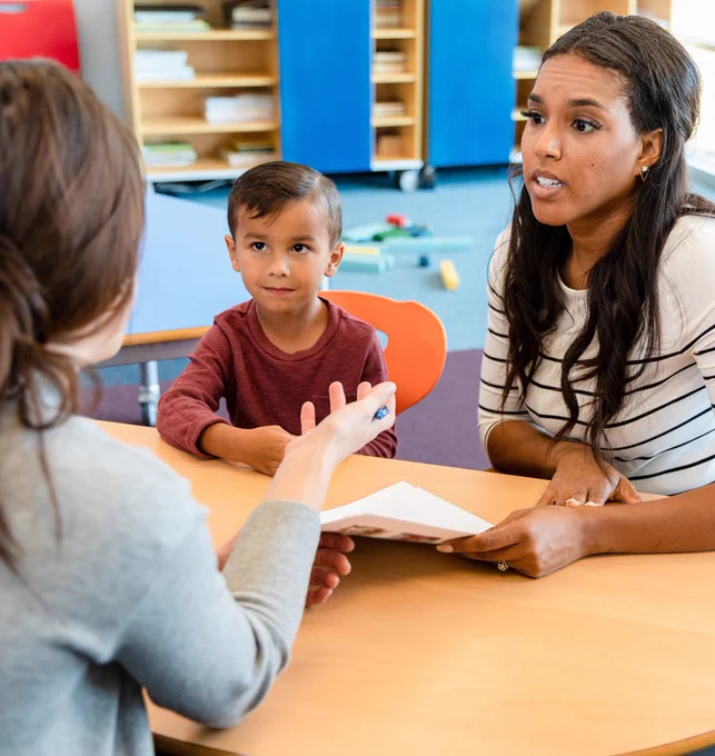 A parent and her son sitting at a table meeting with the son's teacher, who is facing away from the camera in the foreground.  
