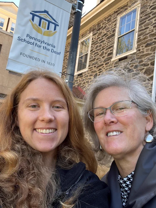 Two women take a selfie picture together outside with a banner behind them that reads Pennsylvania School for the Deaf.