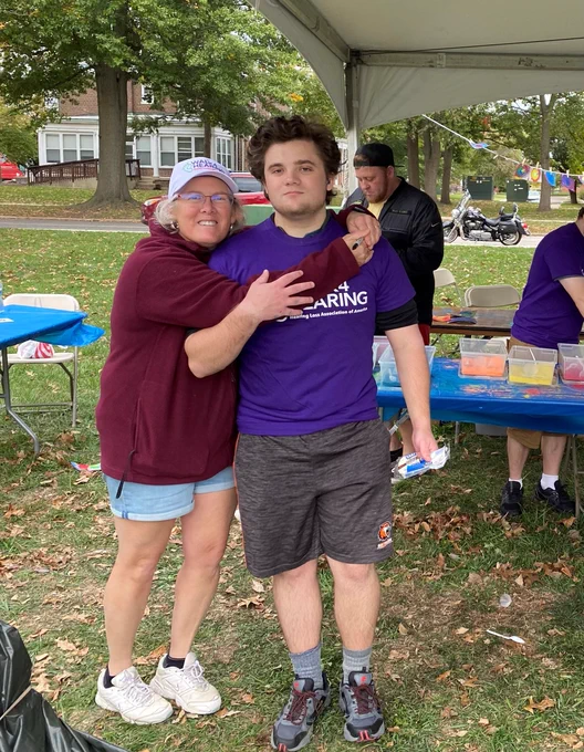 A woman and a man stand together outside. The man is wearing a purple T-shirt and black shorts. The woman is wearing a white hat, dark red hoodie, and blue shorts.