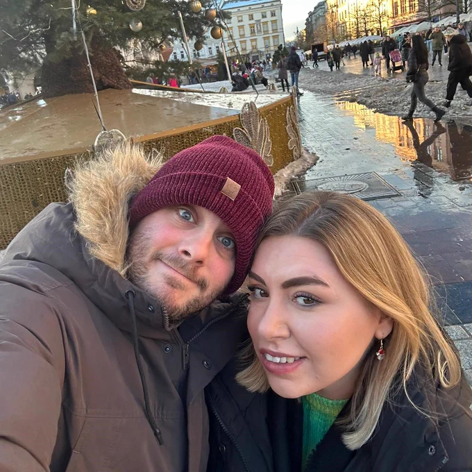 A photo of a man and woman on holiday. The man is wearing a red hat and a coat and the woman is wearing a coat and a green jumper.
