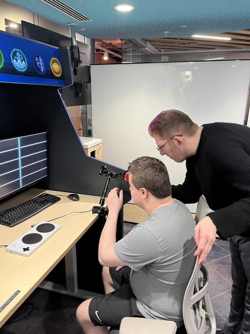 One man using head switch being assisted by another person in front of gaming console
