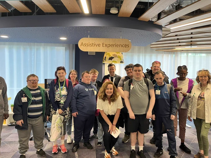 Group of people smiling look at camera with Assistive Experience above on a wooden sign