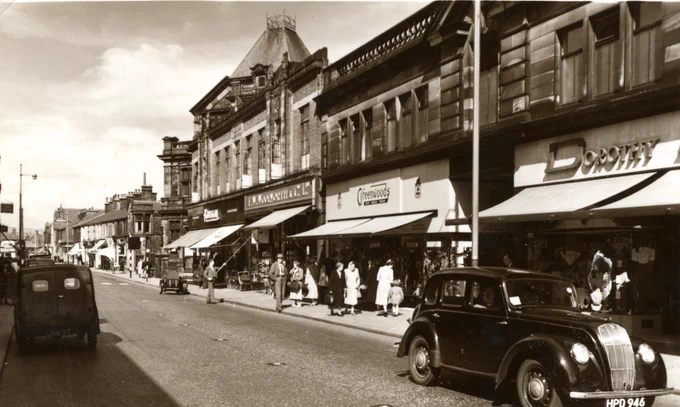 Black and white photo of Scotland Road, Nelson (c.1950) showing shops, cars and people on the pavement