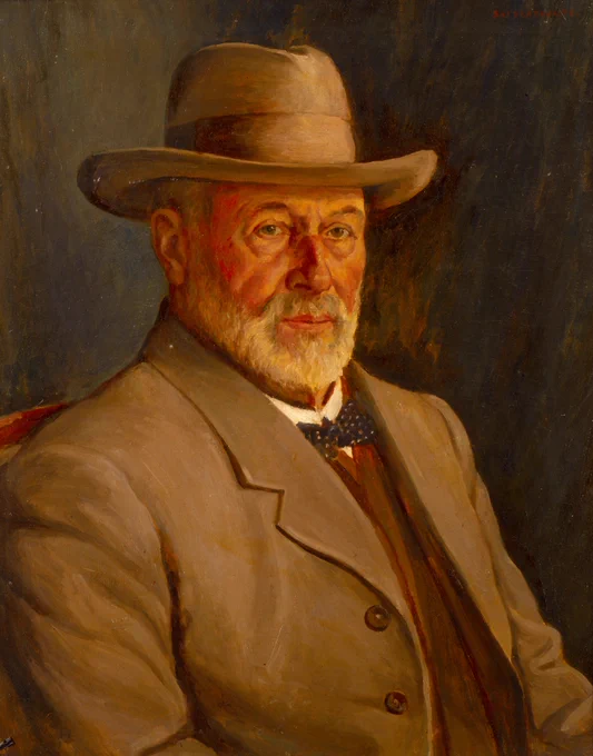 A portrait of a bearded, grey-haired white man wearing a taupe wide brimmed hat and jacket and a spotted bow tie, with neutral expression.
