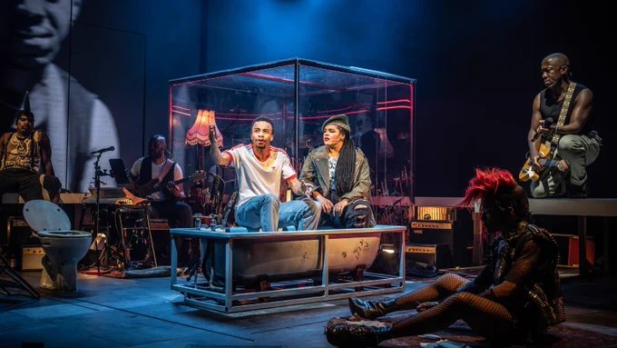 Two people sit in a bathub: an average height, slim, Afro-Caribbean man wearing a red and white top and jeans, and a mixed race girl with long braids, wearing a green khaki jacket and a beret. They are on a stage with other figures surrounding them; one of the figures' face is projected onto a video wall behind them. 