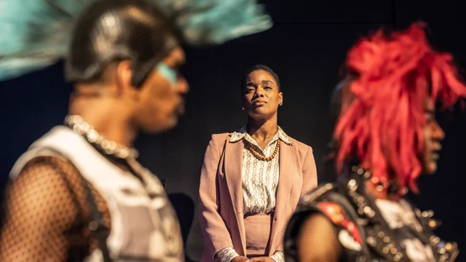 A Black woman of average height with short hair, wearing a peach coloured suit, stands between a Black man and tall Black woman in the foreground, with their hair styled into mohawks, wearing black mesh shirts and sleeveless jackets. 